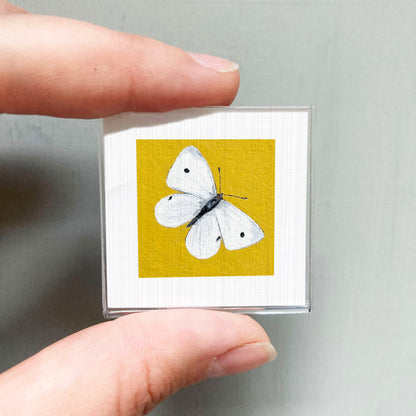 A hand is holding a small square with a white butterfly painted on it against a yellow background.
