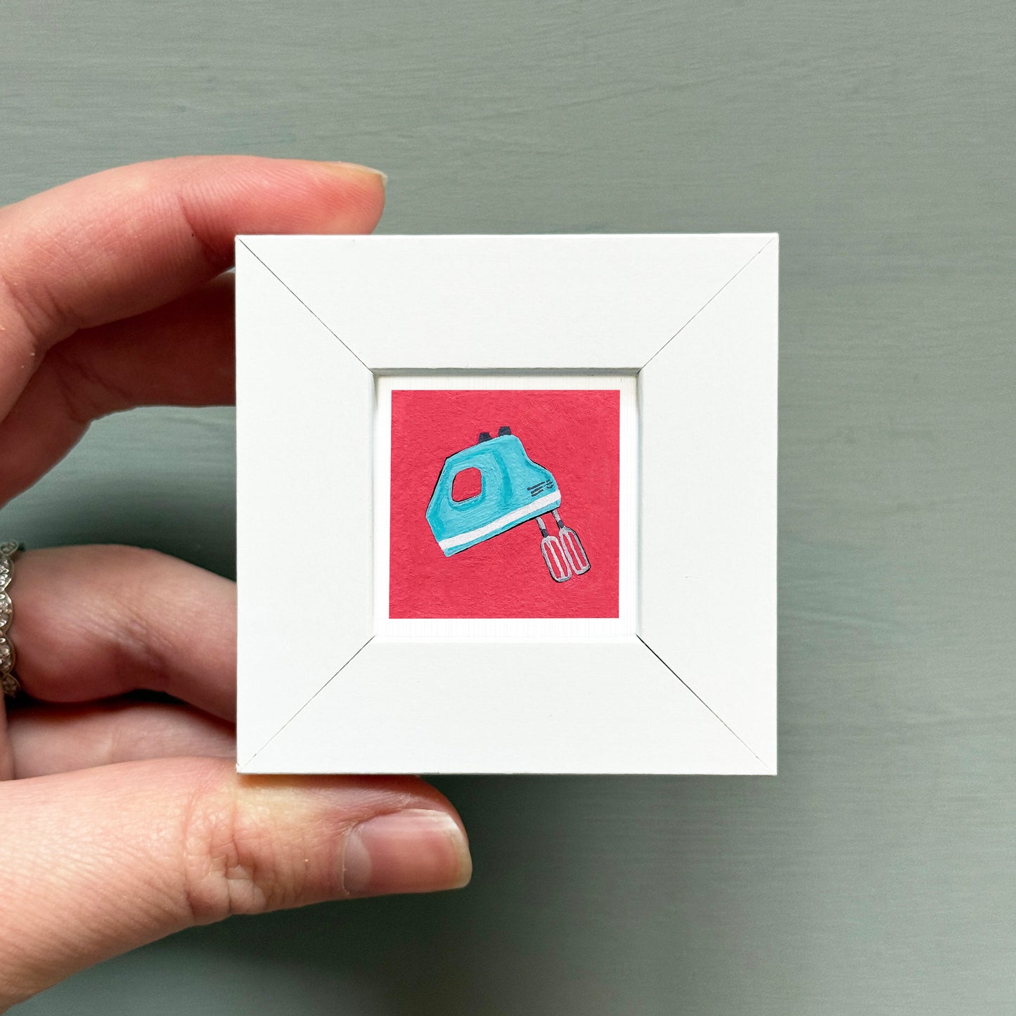 A hand is holding a small white square frame containing an illustration of a blue and white blender.