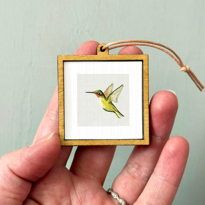 A person's hand holding a small wooden frame containing a framed image of a hummingbird.