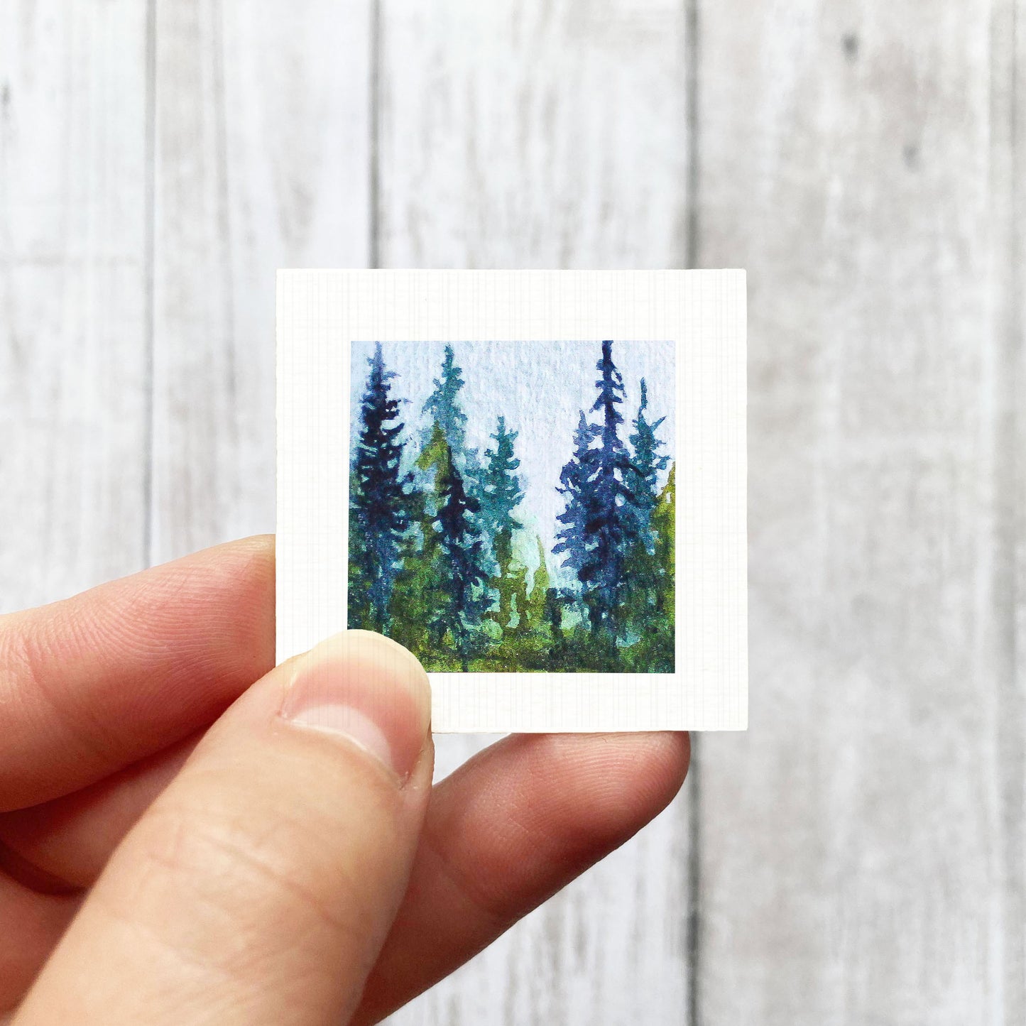 A hand is holding a small square painting of a forest scene with tall trees in shades of blue and green.
