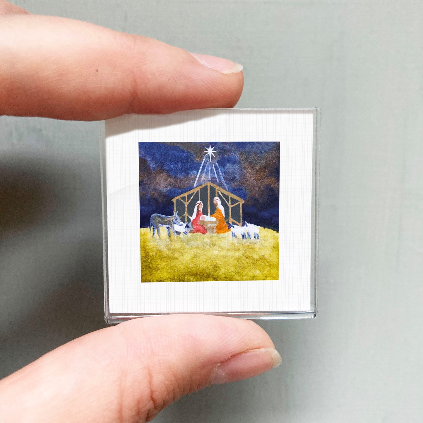 A person's hand is holding a small square picture depicting a nativity scene with a man, a woman, and a baby in a manger, surrounded by animals.