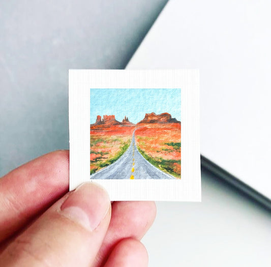 A hand is holding a small square picture depicting a desert landscape with a winding road leading towards a mountain range.