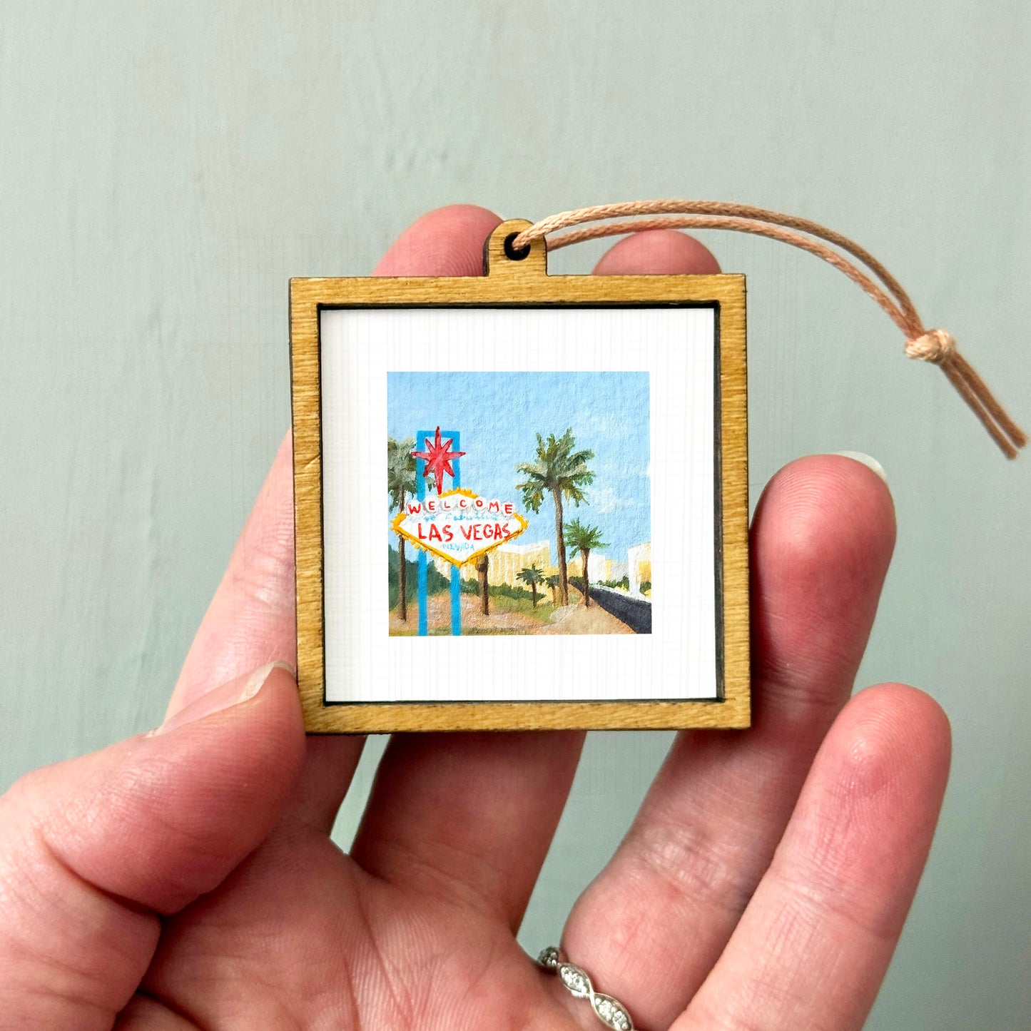 A person's hand holding a small wooden frame containing a framed painting of a Las Vegas street sign.