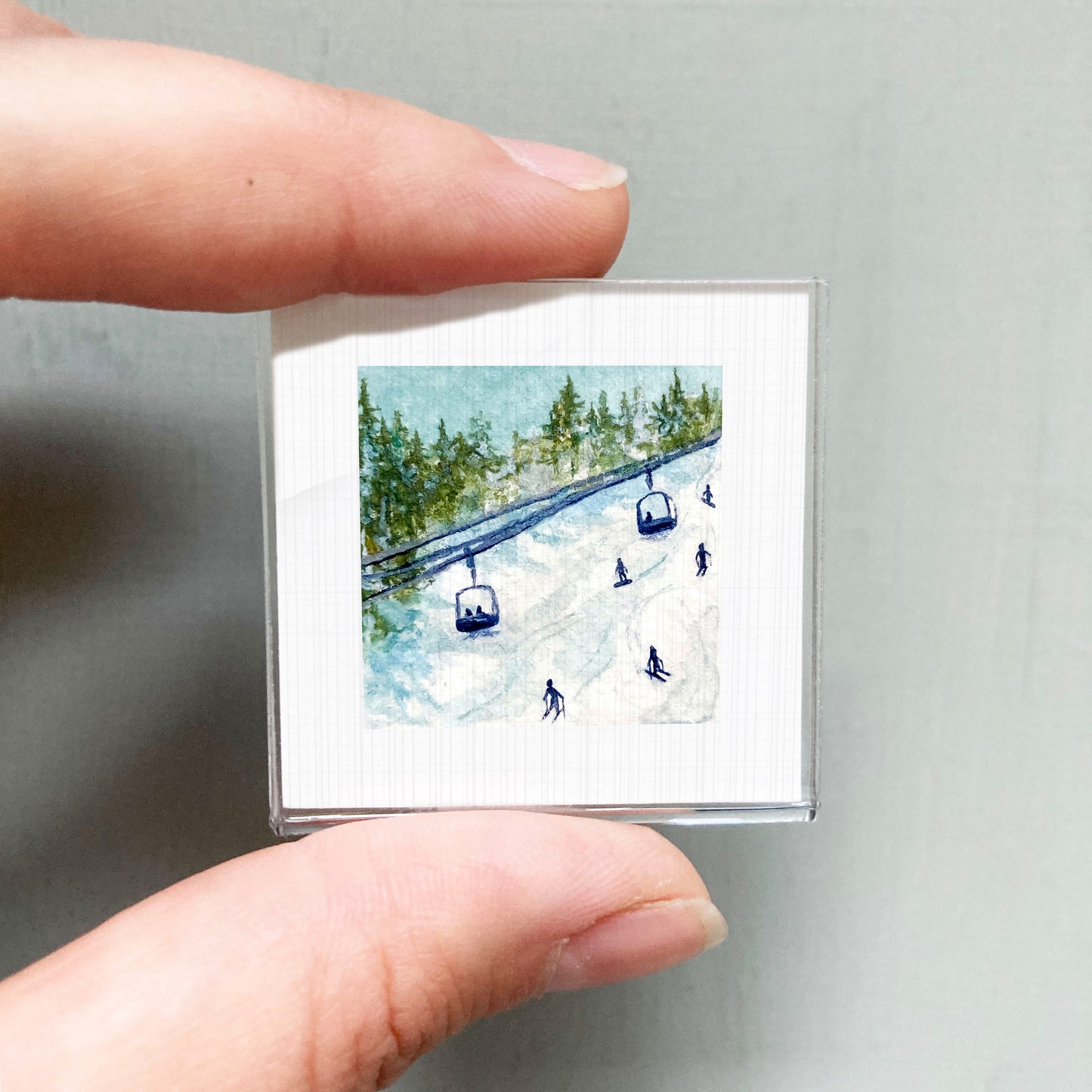 A person's hand holding a small square picture depicting a snowy ski slope with ski lifts and people skiing.