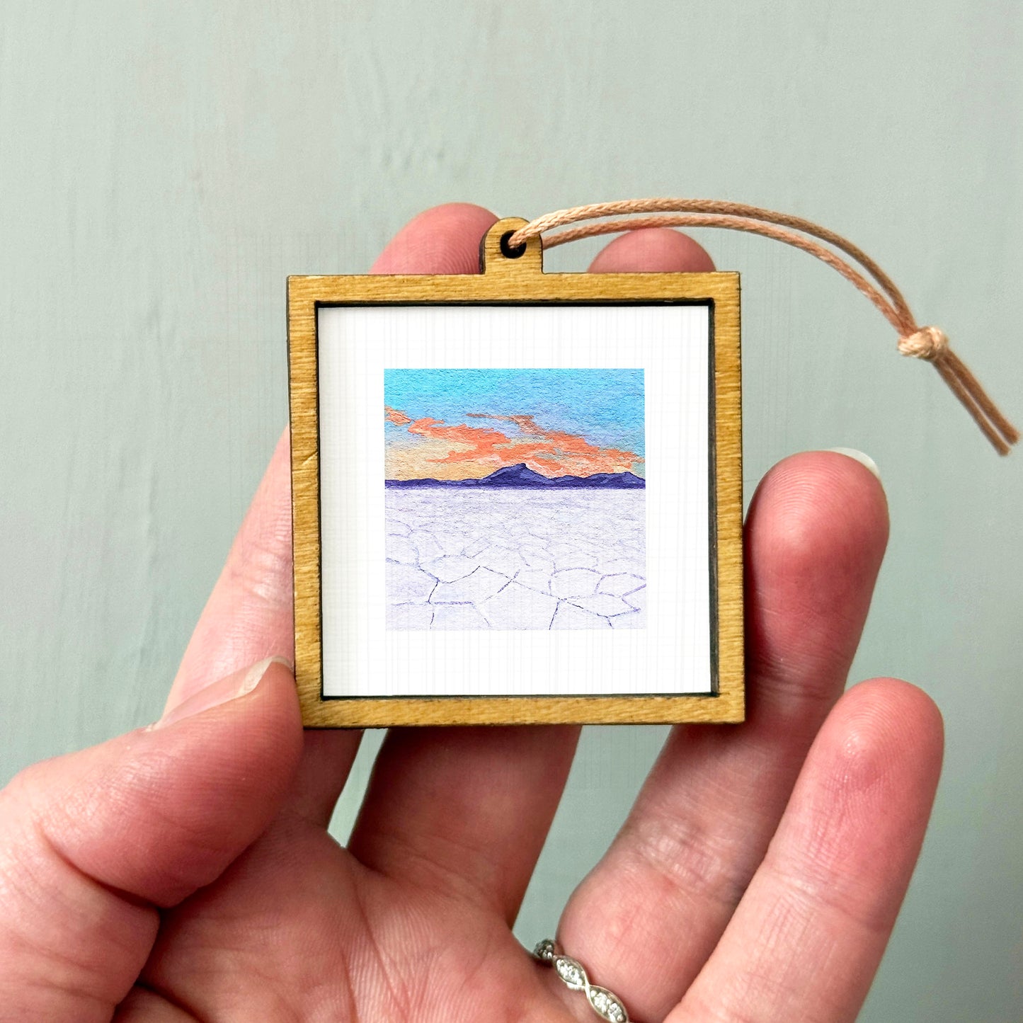 A hand holding a small wooden frame containing a framed landscape painting depicting a desert scene with mountains in the background.