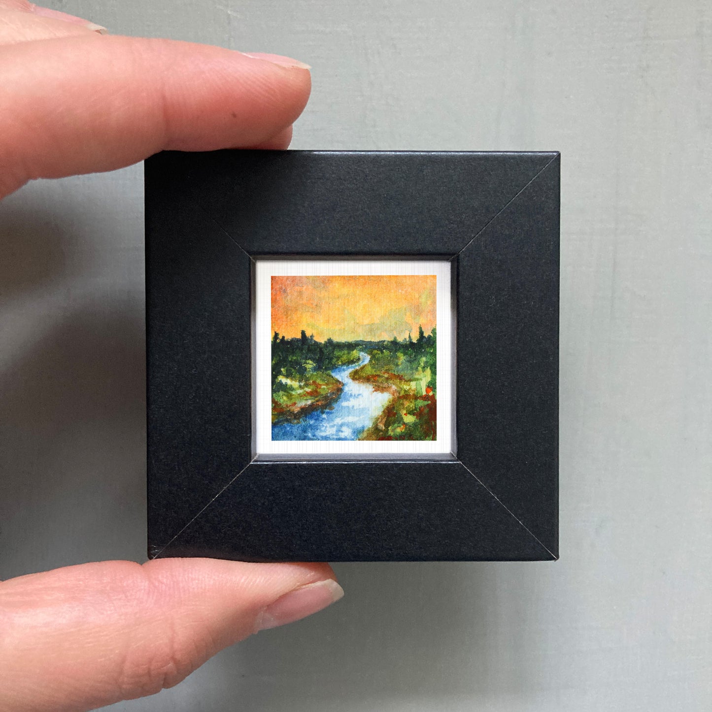 A person's hand holding a small, square-shaped black frame containing a miniature painting of a river and trees.