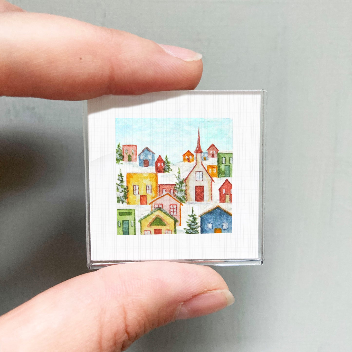 A hand is holding a small square picture depicting a colorful, whimsical winter village scene with houses, trees, and a church spire.