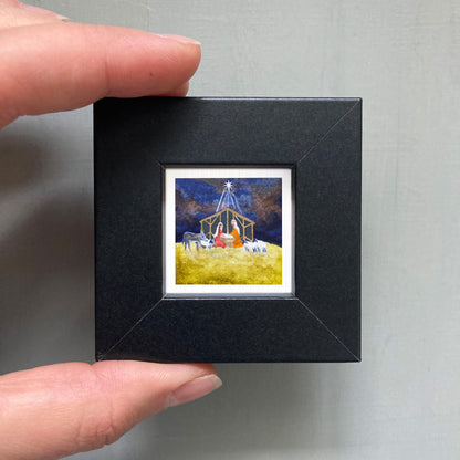 A hand is holding a small black frame containing a framed painting of a nativity scene.