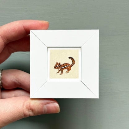 A person's hand is holding a small white square frame containing a painting of a chipmunk.