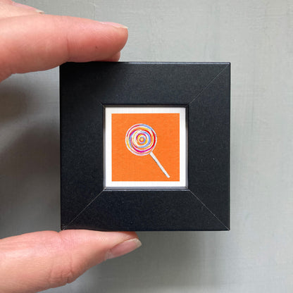 A hand is holding a small black square frame containing a vibrant, colorful lollipop illustration.