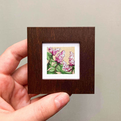 A hand is holding a small wooden frame containing a framed painting of pink flowers.