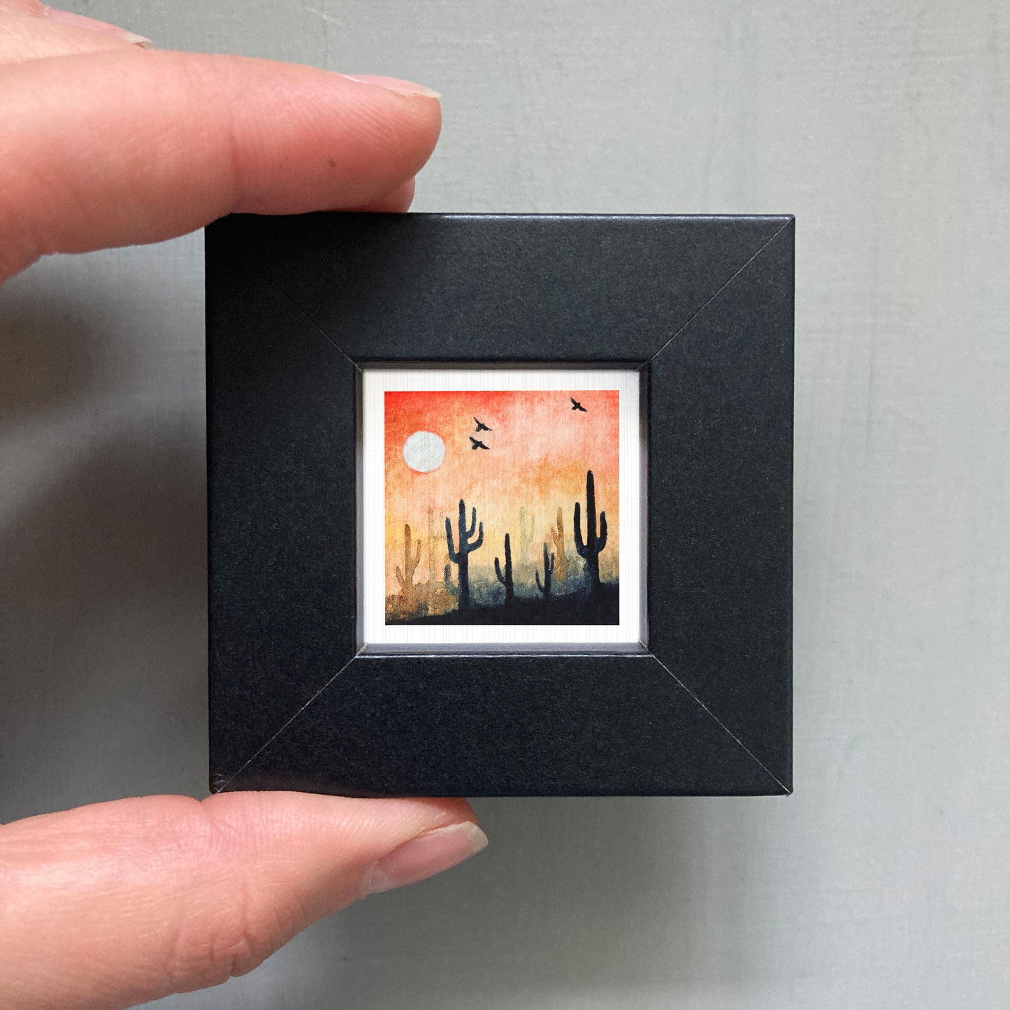 A hand is holding a small black frame containing a framed painting of a desert landscape with cacti and a large white moon in the sky.