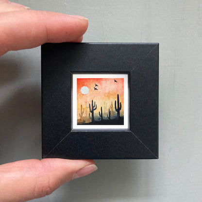 A hand is holding a small black frame containing a framed painting of a desert landscape with cacti and a large white moon in the sky.