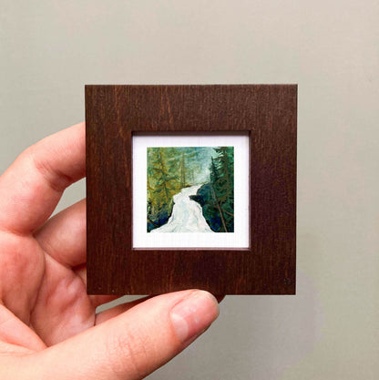 A hand is holding a small wooden frame containing a painting of a forest scene with a winding path.