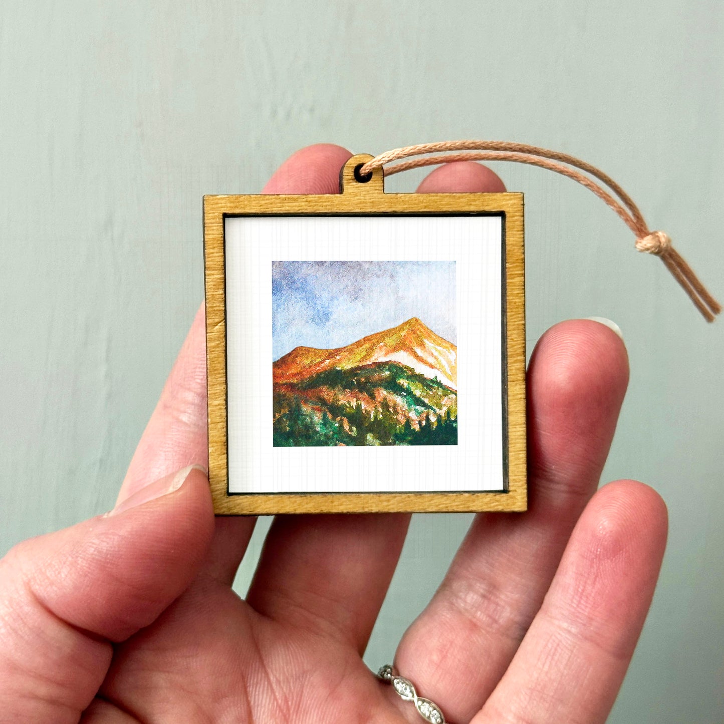 A hand holding a small wooden frame containing a watercolor painting of a mountain landscape.