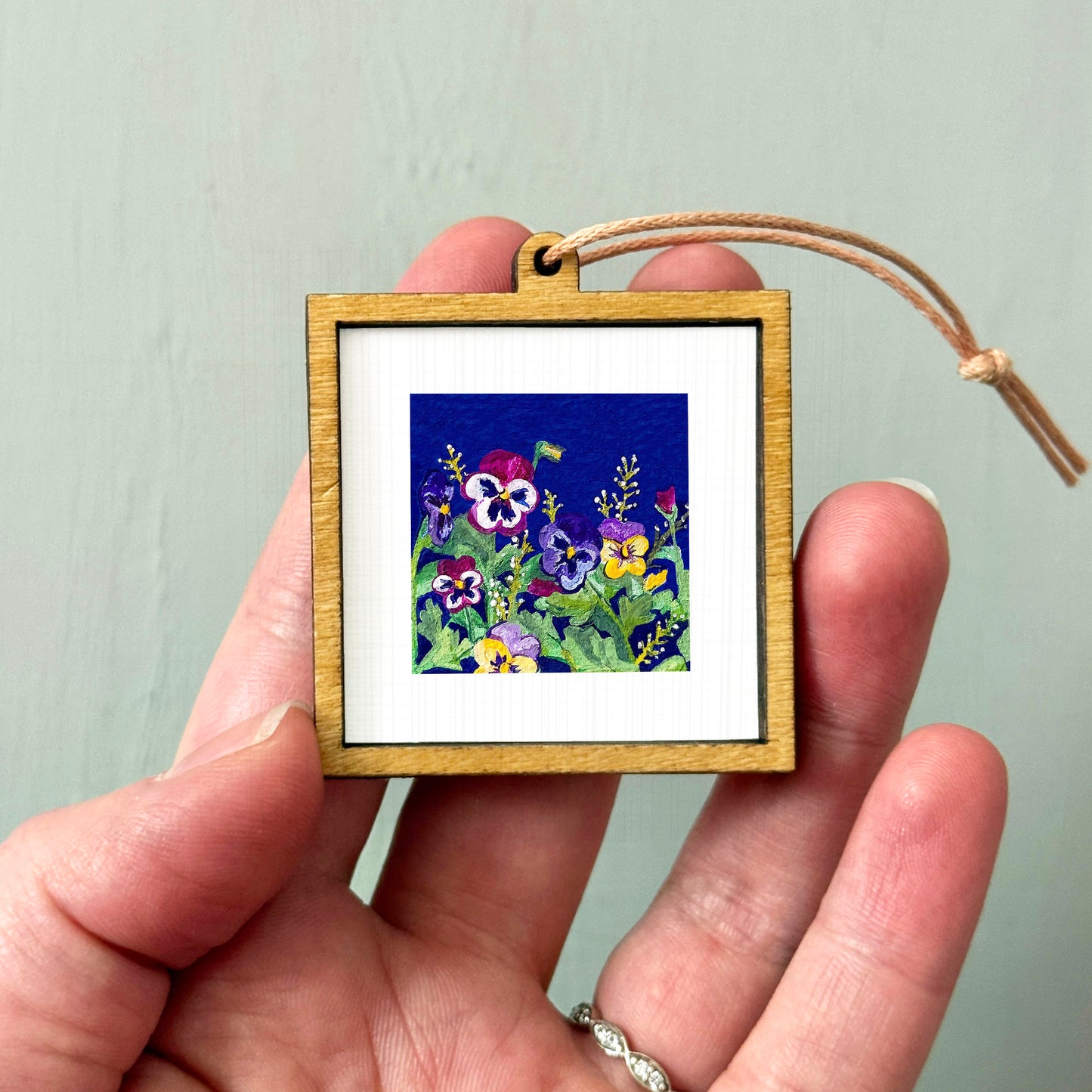 A hand holding a small wooden frame containing a colorful painting of flowers.