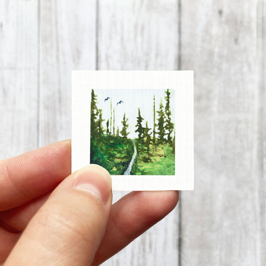 A hand is holding a small square-shaped piece of paper with a painting of a forest scene on it.