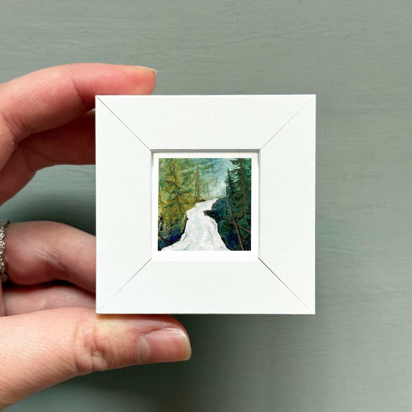 A hand is holding a small white square frame containing a painting of a forest scene with a winding river.