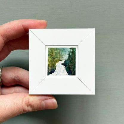 A hand is holding a small white square frame containing a painting of a forest scene with a winding river.