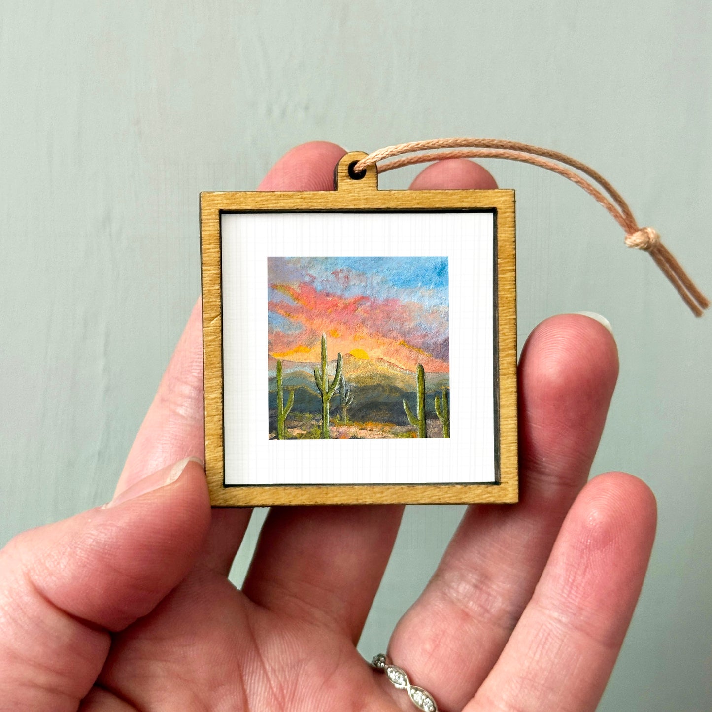 A hand holding a small wooden frame containing a painting of a desert landscape with cacti.