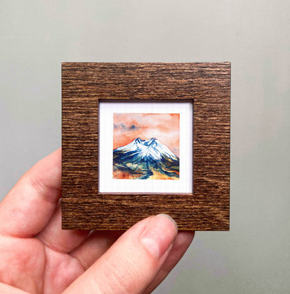 A hand is holding a small wooden frame containing a watercolor painting of a mountain landscape.