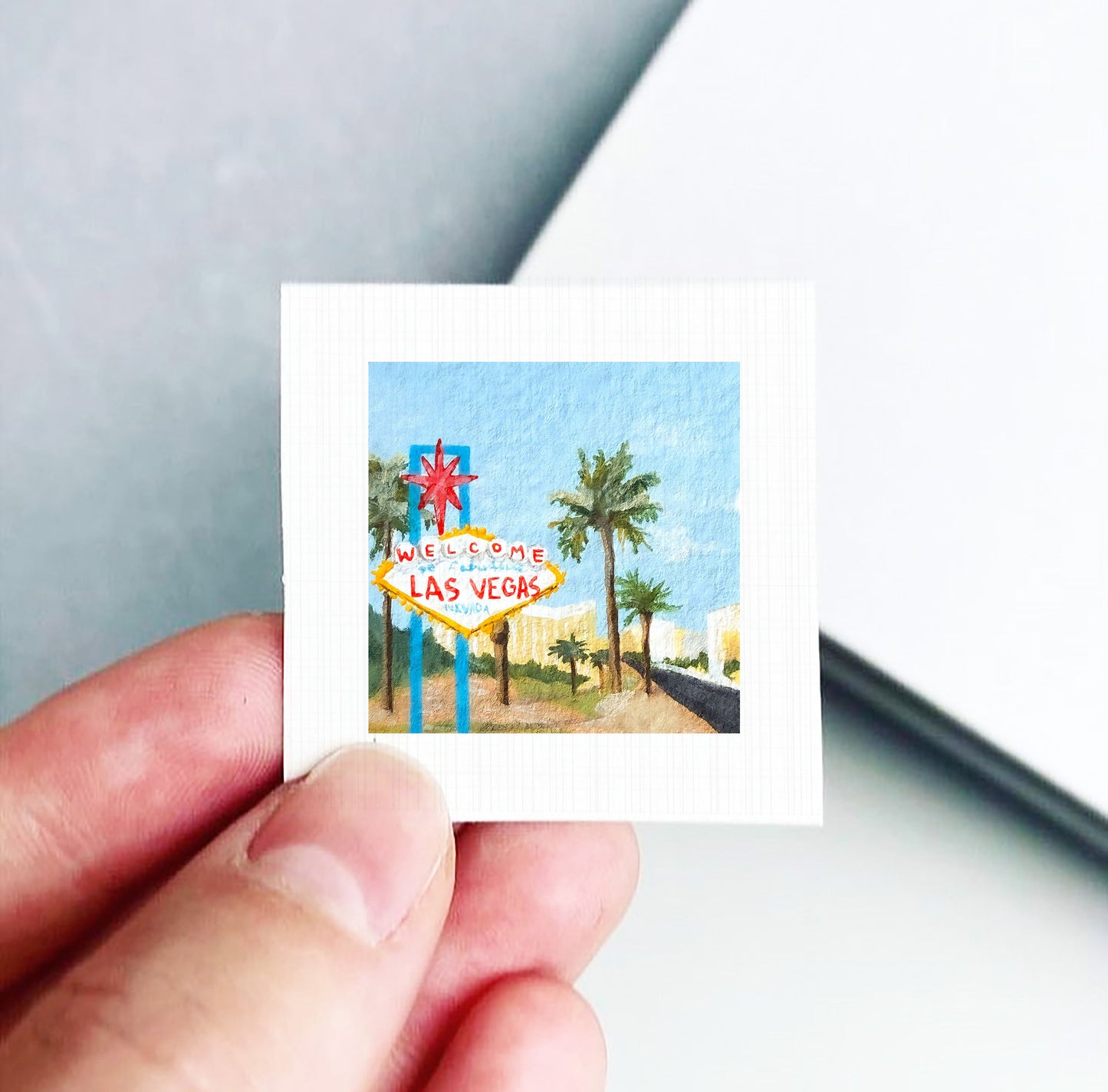 A hand is holding a small square-shaped piece of paper with a painting of the Las Vegas sign.