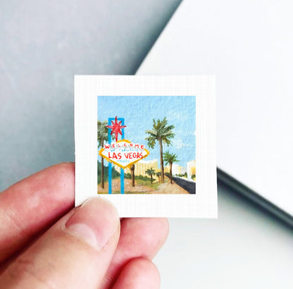 A hand is holding a small square-shaped piece of paper with a painting of the Las Vegas sign.