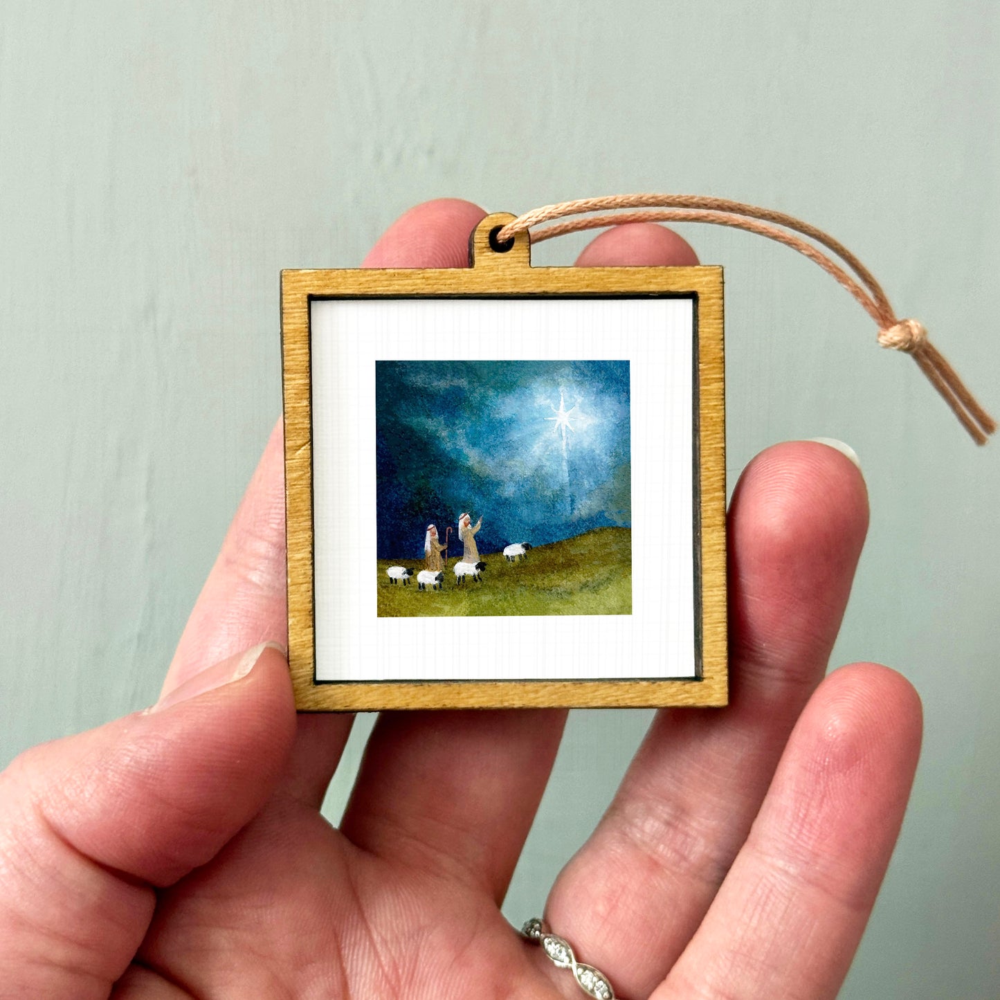 A hand holding a small wooden frame containing a painting of a nativity scene with two figures and sheep.