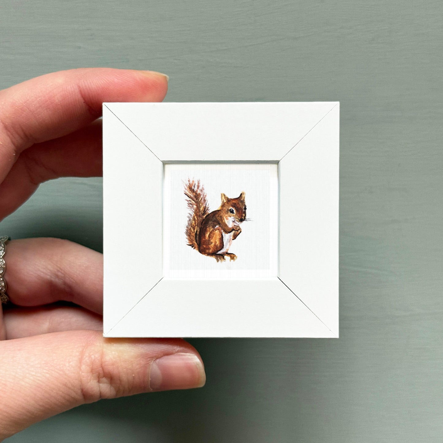 A hand is holding a small white square frame containing a painting of a squirrel.