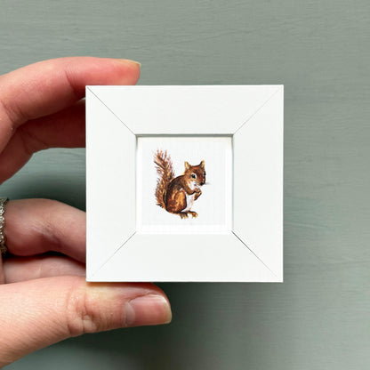 A hand is holding a small white square frame containing a painting of a squirrel.
