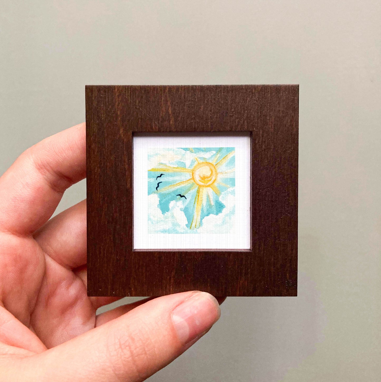A hand is holding a small wooden frame containing a framed painting of a sunny sky with birds.