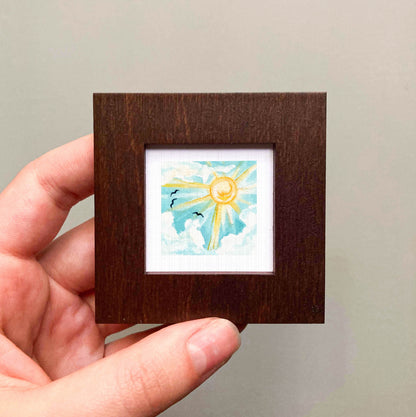 A hand is holding a small wooden frame containing a framed painting of a sunny sky with birds.