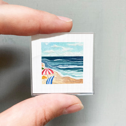 A person's hand holding a small square picture depicting a beach scene with umbrellas and the ocean.