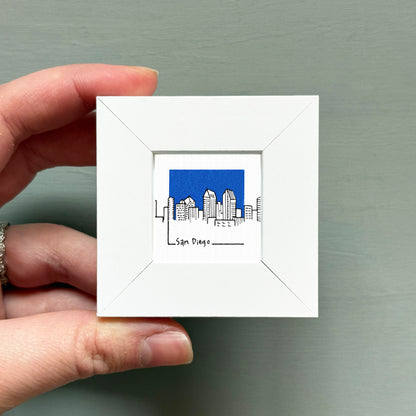 A hand is holding a small white square frame containing a miniature illustration of the San Diego skyline.