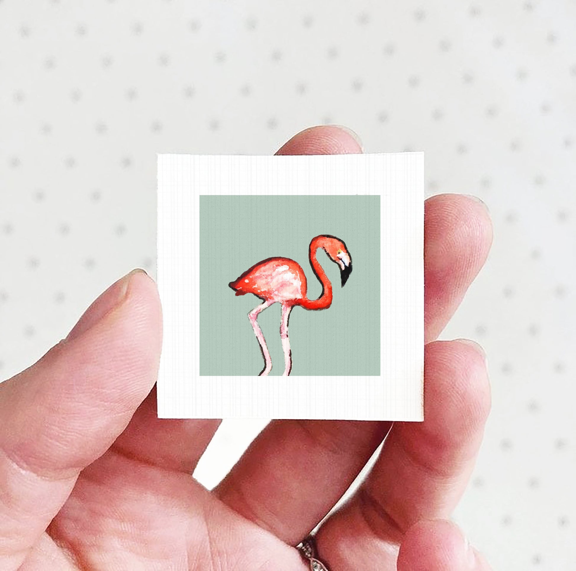 A hand holding a small square picture of a flamingo against a light green background.
