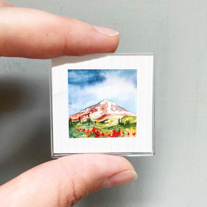 A person's hand holding a small, square-shaped picture of a mountain landscape with a colorful, vibrant scene.