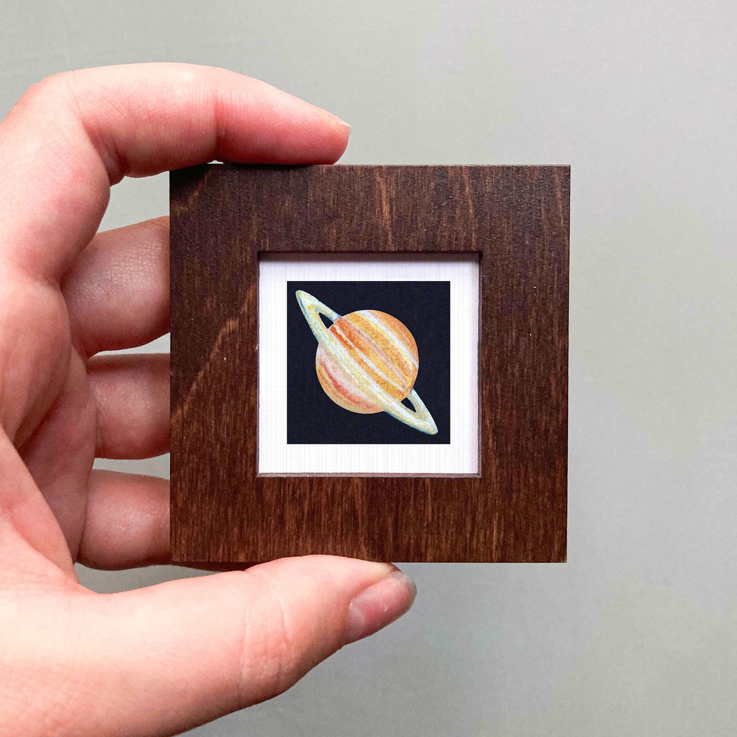 A hand is holding a small wooden frame containing a framed image of a planet.