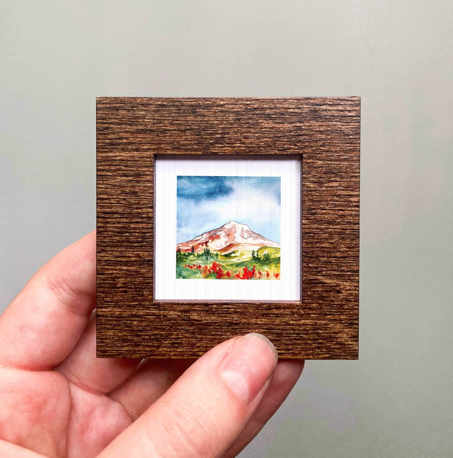 A hand holding a small, square-shaped wooden frame containing a watercolor painting of a mountain landscape.