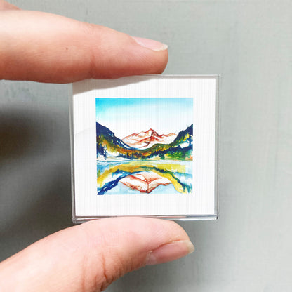 A person's hand holding a small, square-shaped picture of a mountain landscape with a lake and mountains in the background.