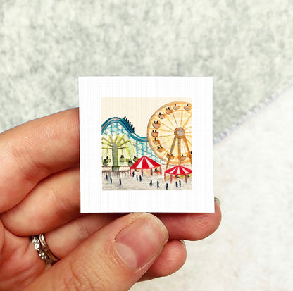 A hand holding a small square artwork depicting a colorful amusement park scene with a roller coaster and other rides.