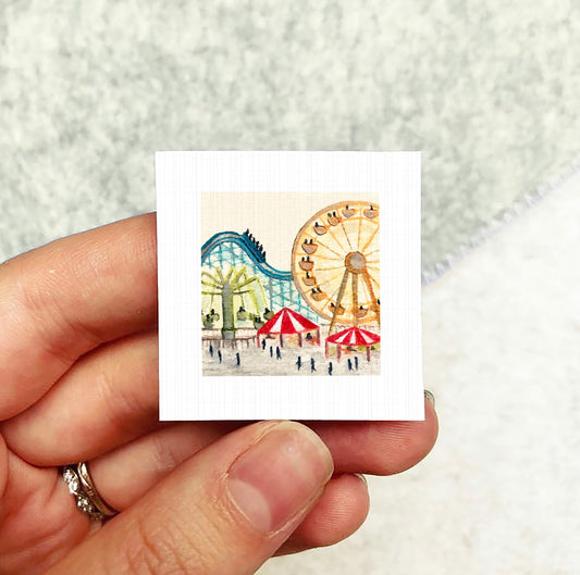 A hand holding a small square artwork depicting a colorful amusement park scene with a roller coaster and other rides.