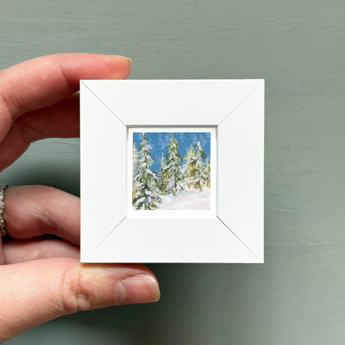 A hand is holding a small white square frame containing a painting of snow-covered pine trees.