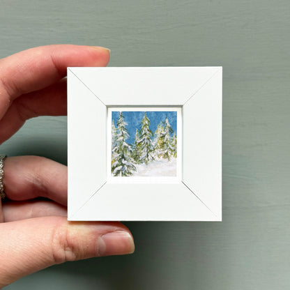 A hand is holding a small white square frame containing a painting of snow-covered pine trees.
