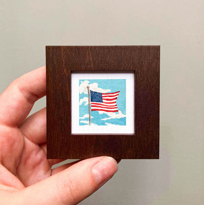 A hand is holding a small wooden frame containing a miniature painting of an American flag on a cloudy sky.