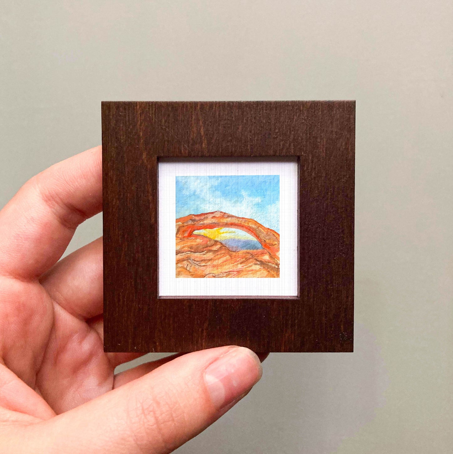 A hand is holding a small wooden frame containing a watercolor painting of a desert landscape with a rainbow arching over the scene.