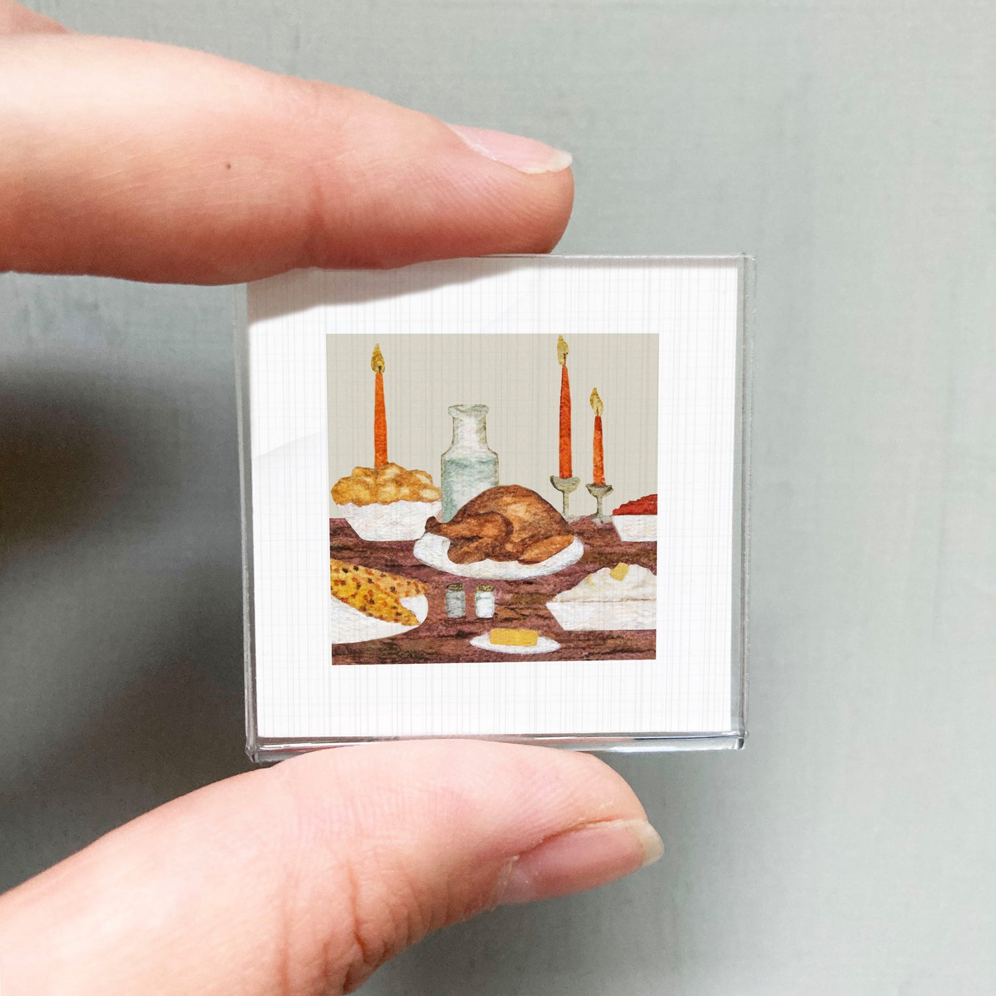 A person's hand is holding a small square picture depicting a table with a roasted turkey, bowls of food, and candles.