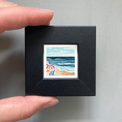 A person's hand holding a small, square-shaped black frame containing a watercolor painting of a beach scene with umbrellas and the ocean.