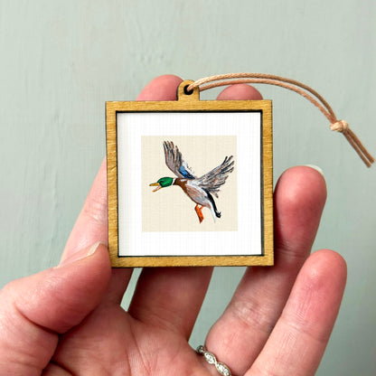 A person's hand holding a small wooden frame containing a framed image of a bird in flight.