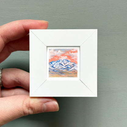 A hand is holding a small white square frame containing a painting of a mountain landscape with a colorful sky.