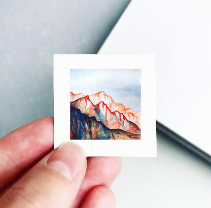 A hand is holding a small square-shaped piece of paper with a painting of a mountain landscape on it.